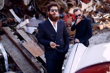 Close up of a bearded mens in black suits and sunglasses smokes cigars near vintage car, on the iron dump background.の写真素材