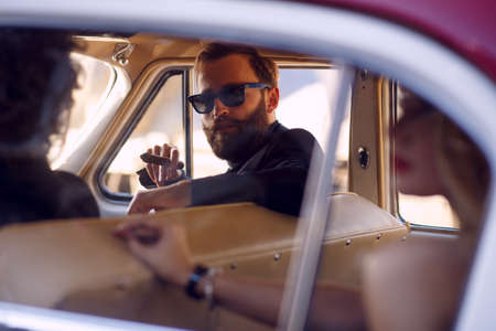 Close up portrait of a stylish bearded man smokes cigar in vintage car. A two men and woman inside of a vintage car.の写真素材