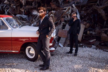 A two bearded men in a classic suits, sunglasses, smoking cigars, on iron dump background. Gentlemen's club.の写真素材