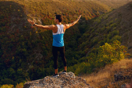 Back of a athlete, muscular, fit, abs, young man doing stretching exercises with hands through the parts, before workout outside on forest, mountain background. Summer landscape. Copy space.の写真素材