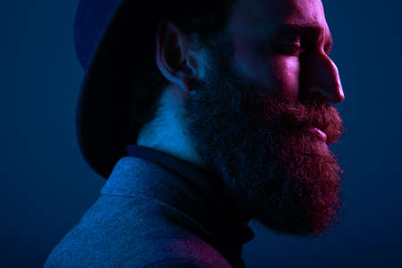 Portrait of a bearded man in hat and suit, with close eyes and eyeglasses posing in profile in studio, keeping palms pressed together, isolated on dark blue background. Horizontal view.の写真素材