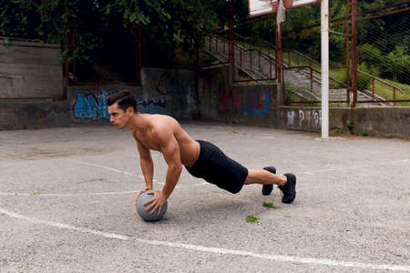 A fit, strong, muscular young man shirtless holding a bascketball, poses outside in urban court. Sport concept.の写真素材