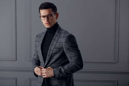 A fit man in gray suit with black turtleneck,black stylish eyeglasses looks confident at the camera.Perfect model of a man of affairs.Horizontal view on gray wall background.の写真素材