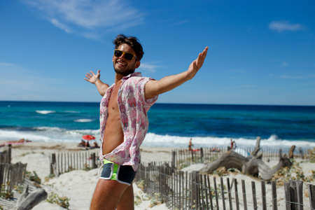 A happy sexy young man posing with raised hands on the beach, dressed in a swimwear and summer shirt, isolated on beach, ocean background. Horizontal view Corsica island, France.の写真素材
