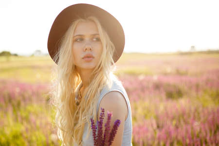 Beautiful bright soft girl with long blond curly hair wearing a hat with large fields in the summer pink sundress standing near a pink floral field with violet flowers.の写真素材