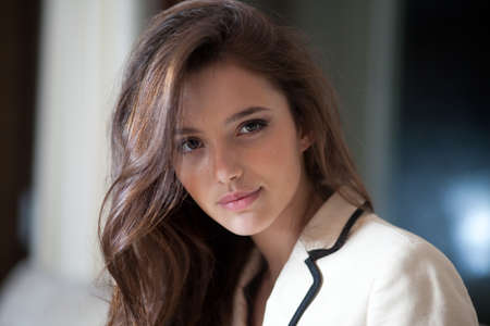 Beautiful portrait of a young elegant woman with long hair, wearing in delicate suit, looking confident at camera, blurry room background.の写真素材