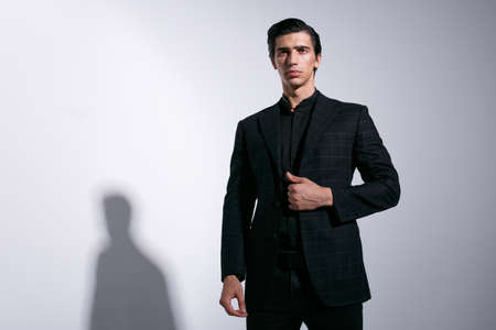 Young sexy handsome male model dressed in black suits on white studio background with shade on the wall. Horizontal indoors shot.の写真素材