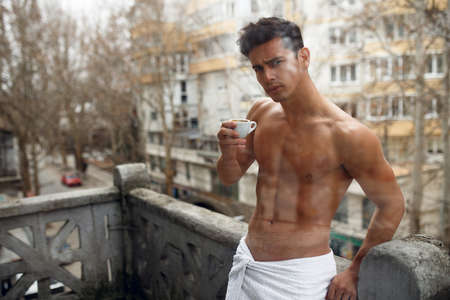 Sexy brunette young naked man holding in hands a cup of coffee, posing to the balcony, on street, urban, city background. Horizontal view.の写真素材