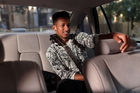 Young man multi race sitting on the back seat in the car. A singer holding a guitar while traveling...