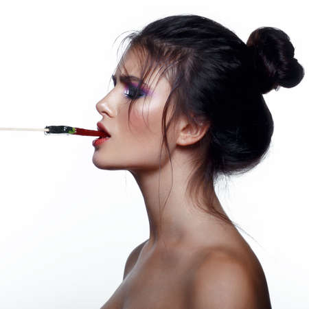 Beauty portrait of an excited brown haired woman with makeup eating lollipop and looking in profile,...