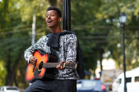 Profile of a happy young musician played on guitar, standing near street lamp sing a song in sunny...