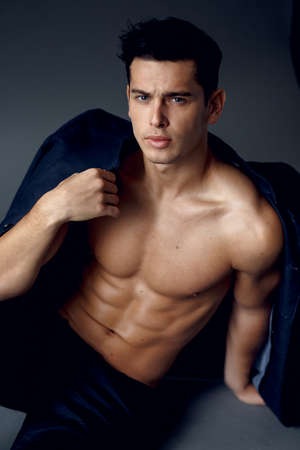 A young, handsome, brunette sexy man with jacket on his shoulders, with naked torso, posing sitting on grey background, modern guy, lifestyle people concept. Vertical view.の写真素材