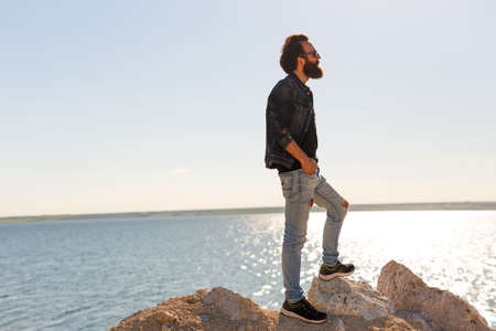 Side view of a stylish bearded hipster boy in sunglasses posing near a calm sea during a wonderful journey around the world. Shoot from the side.の写真素材