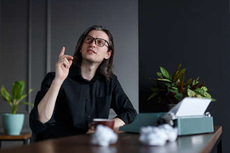 Portrait of a man in black shirt, with long hair, raised his finger up show have a ideea, seated at a table near typewriter in his office. A writer searching a inspiration, over gray background.の写真素材