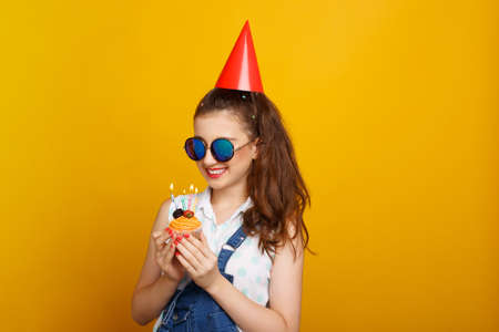 Girl in sunglasses holding in hands a cupcake with candles. Teenager with birthday cupcake on yellow...