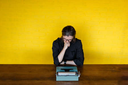 In search of inspiration. Thoughtful mature man holding hand under the chin, looking on down on a typewriter while sitting at his working place in office, over yellow background.の写真素材