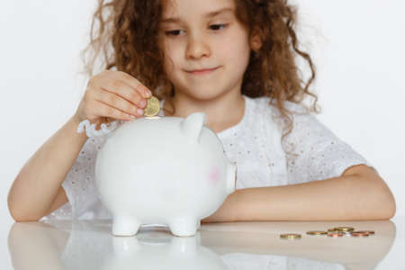 Closeup portrait of a cute curly girl in white putting coin into piggy bank, beside a lot of coins, over white background.の写真素材