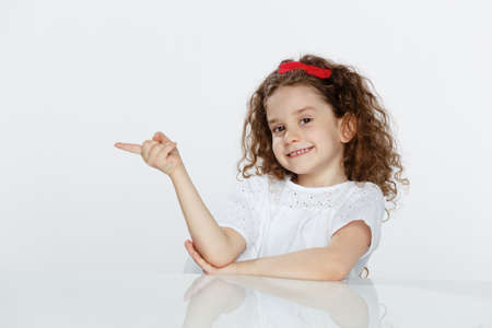 Portrait of a little adorable curly girl, seated at table, showing with finger on lateral direction, over white background. Copy space.の写真素材