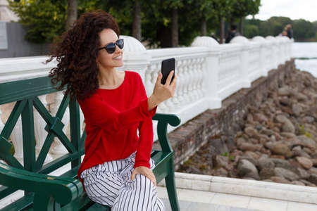 Stylish beautiful curly woman in sunglasses doing selfie on a smartphone, seated on a green bench in the park. Background in summer port. Place for text. Horizontal view.の写真素材