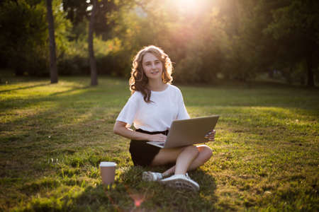 Girl with laptop sitting on the green grass and smiling, looking at camera. Educational concept. Girl working on a laptope outside.の写真素材