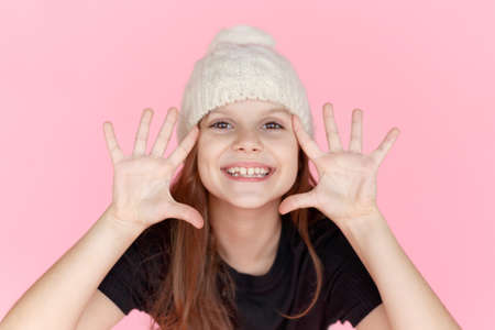 Frontal portrait of a cute smiling little girl in wool cap with open palms near face, looking at...