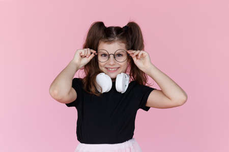 Frontal portrait of a cute brunette little girl with two tails, in black shirt and white headphones, holds his hands on his eyeglasses, over pink background. Horizontal view.の写真素材