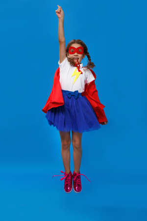 Frontal portat of a very excited little girl dressed like superhero jumping alongside the blue wall....