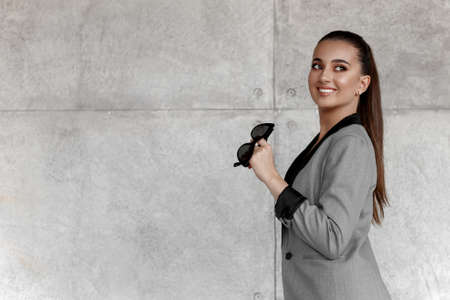 Young smiling busines woman in grey jacket with black sunglasses in hand, looking on the side, with...