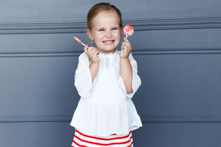 Cheerful little girl holding lollipop, smiling and looking at camera, isolated on grey neoclassic background. Copy space.の写真素材