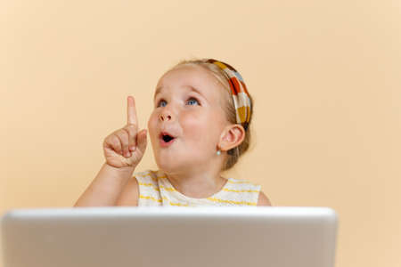 Kid point finger up isolated beige. Little girl with laptop pointing upwards, having idea or...