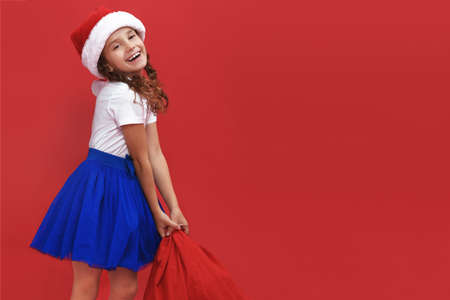 Adorable little smiling girl wearing santa hat carrying gift bag isolated on red background. Space for text.の写真素材