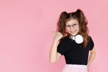 Little girl in eyeglasses, white headphones, looking at camera and smiling, pointing something, over pink background.の写真素材