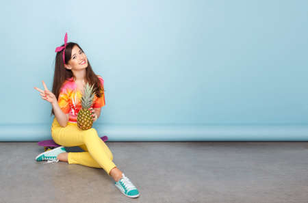 Cheerful teen girl posing in studio in colorful clothes, seated on skateboard, holding un pineapple, show victory sign.の写真素材