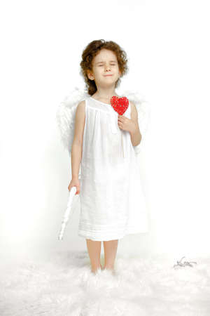Dreaming little girl with curly hair, in white dress and wings, over white background. Angel holding a red heart.の写真素材