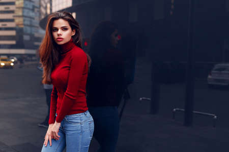 Pretty sexy girl in a fashionable red sweater in blue jeans posing on the street near a wall.