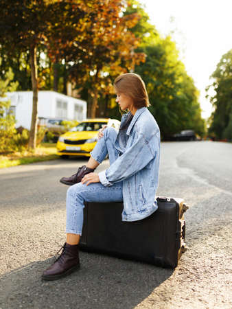 Profile portrait pensive girl sits on her suitcase on middle road, waiting a transportation, in sunset background.の写真素材