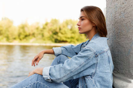 Profile of a serious single pensive teen girl, dressed in denim, seated outside near river, looking away.の写真素材