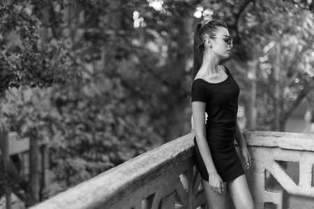 Sensual lady, sublime and fit body, in black dress posing in sunglasses, near the balcony. black and...