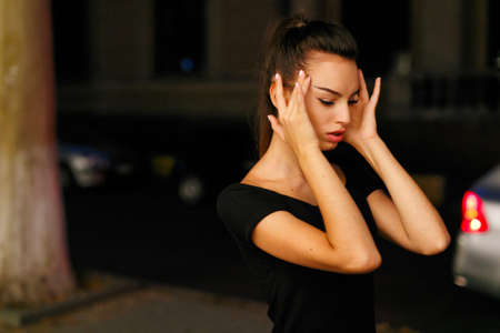 Young girl in profile, holds her head in her hands, suffers from headache, sits in the street, at...