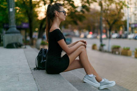 Full length portrait. Lonely girl sits on the stairs in the city, in a black dress with sunglasses,...