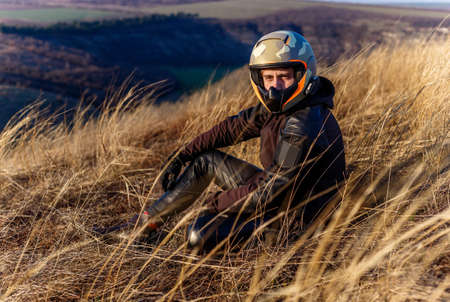 Portrait of a young sportsman, a biker in helmet and leather jacket looking at camera, seated on a dry field.の写真素材