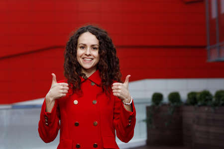 Smiling pretty brunette curly young woman in red suit, showing thumbs up isolated on the red building background.の写真素材