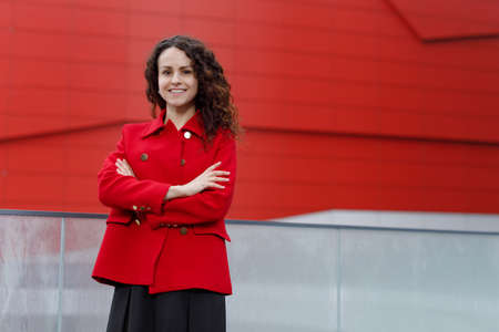 Cheerful young woman with curly hair, with a beautiful smile, keeps her hands crossed, dressed in a red jacket. Picture of a business woman, looks confidently at the camera.の写真素材