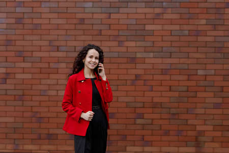 Profile portrait of woman in red, hair in wind, talking on the smartphone while walking outside, near brick wall. woman happy using mobile phone app for conversation. Space for text.の写真素材