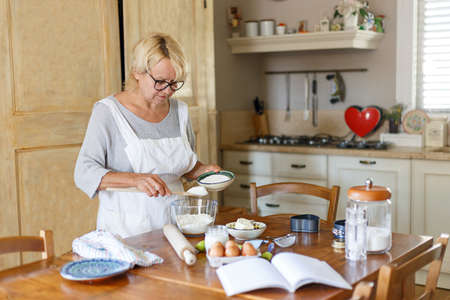 Horizontal view. Beautiful grandmother in white apron, cooking on the kitchen, has on the table ingredients to bake.の写真素材