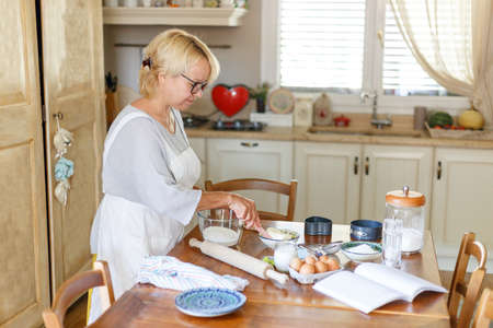 Profile of beautiful peaceful grandmother in white apron, cooking on the kitchen, has on the table ingredients to bake.の写真素材