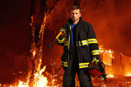Firefighter with sad equipment, holds the helmet in his hand, which stands against the background of a forest in flames.の写真素材