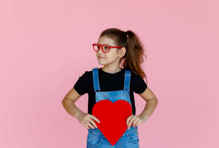 Cheerful cute little girl in red eyeglasses, holding a big red valentines heart, looking away, over pink background.の写真素材