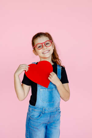 Happy little girl in red eyeglasses, wear in black shirt, holding a big red valentines heart, over pink background.の写真素材