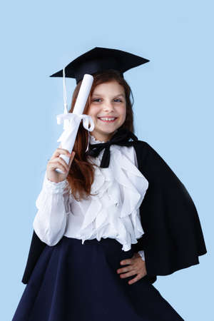 Portrait of a cheerful little girl in graduation cap standing and showing a success moment, over blue background.の写真素材